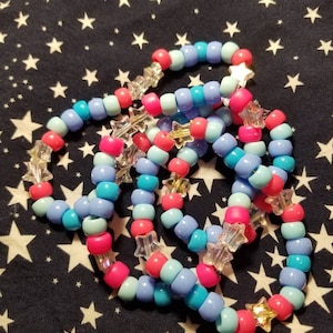 May include: A set of colorful beaded bracelets with star-shaped charms. The bracelets are made with light blue, turquoise, and pink beads.