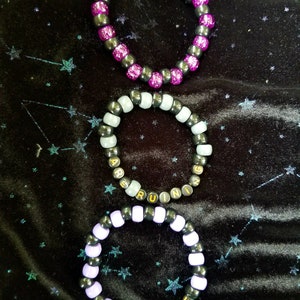 May include: Three beaded bracelets with black beads and gold lettering. The top bracelet has pink beads and says "I LOVE GIRLS". The middle bracelet has white beads and says "ARE RUINING". The bottom bracelet has purple beads and says "MY LIFE".