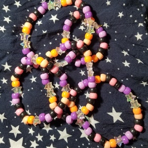 May include: A set of four colourful friendship bracelets with star-shaped beads. The bracelets are made with black, orange, pink, and purple beads.