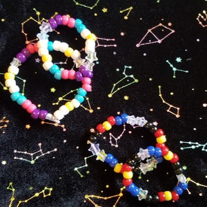 May include: Three colorful beaded bracelets with star-shaped charms. The bracelets are made with small, round beads in various colors, including purple, pink, blue, yellow, white, red, and black. The bracelets are arranged on a black background with a constellation pattern.
