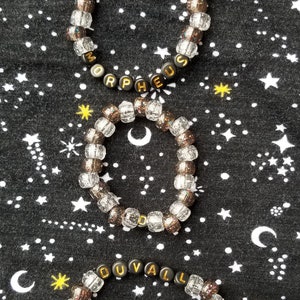 Puede incluir: Tres pulseras de cuentas transparentes con cuentas de letras negras que deletrean los nombres "MORPHEUS", "DUVALL" y "MORPHEUS". Las pulseras están sobre un fondo de tela negra con un patrón de estrellas blancas.