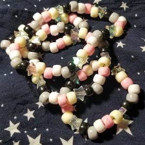 Könnte beinhalten: Ein mehrlagiges Kandi-Armband mit rosa, weißen, grauen und schwarzen Perlen. Jede Perle hat einen sternförmigen Anhänger.