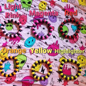 Puede incluir: Seis pulseras de amistad coloridas sobre un fondo rosa con un patrón de caritas sonrientes. Las pulseras están hechas de cuentas negras y de colores. Los colores de las pulseras son rosa claro, magenta, rojo, naranja, amarillo y amarillo fluorescente.