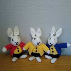 May include: Three knitted white rabbit dolls wearing jackets in red, yellow, and blue. Each doll has black button details and black trousers. The dolls are standing on a wooden surface.