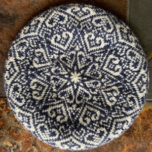 May include: A white and blue knitted beret with a geometric pattern. The beret is circular in shape and has a flat top.