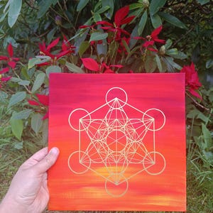 Peut inclure: Une œuvre d'art sur toile carrée avec un dégradé de teintes orange et rouge. Un motif géométrique doré est centré sur la toile. L'œuvre est tenue devant un fond de feuillage vert et de fleurs rouges.