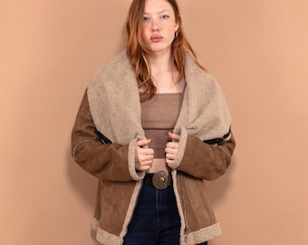 Puede incluir: Una mujer con una chaqueta de gamuza marrón con cuello y forro de piel de oveja marrón claro. Lleva pantalones vaqueros azules y zapatos negros.
