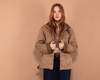 Puede incluir: Una mujer con una chaqueta de gamuza marrón con cuello de piel y pantalones vaqueros azules. Está de pie frente a un fondo marrón claro.