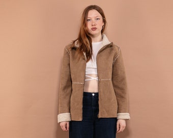Puede incluir: Una mujer con una chaqueta de ante marrón con cuello y ribetes de piel sintética blanca, un top blanco corto y unos vaqueros azules oscuros. Está de pie frente a un fondo marrón claro.