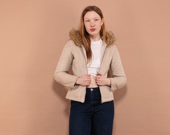Puede incluir: Una mujer con una chaqueta de ante beige con capucha de piel marrón, una camiseta blanca y unos vaqueros azul oscuro. Está de pie frente a un fondo marrón claro.