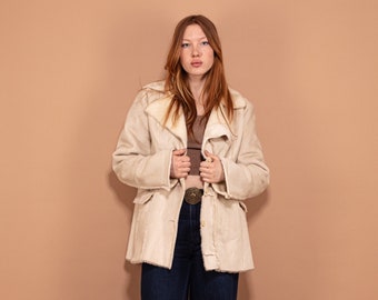 Puede incluir: Una mujer con una chaqueta de piel sintética de color crema, un top marrón y unos vaqueros azules. Está de pie frente a un fondo marrón claro.