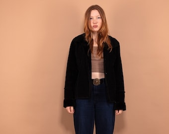 Puede incluir: Chaqueta negra de terciopelo con cuello y mangas largas. Se combina con vaqueros azul oscuro y zapatos negros. Una hebilla dorada añade un toque decorativo. El conjunto se muestra sobre un fondo neutro.