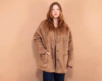 Puede incluir: Una chaqueta de ante marrón con cierre de botones y dos bolsillos de solapa. La chaqueta la lleva puesta una persona con el pelo largo y rojo y está de pie frente a un fondo marrón claro.