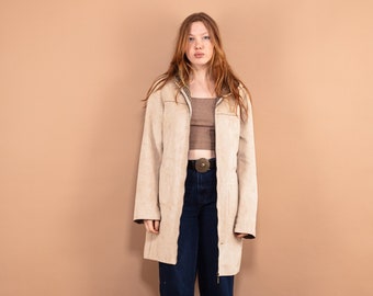 Puede incluir: Una mujer con una chaqueta de gamuza beige con un top marrón debajo. Lleva pantalones vaqueros azules y zapatos negros. El fondo es de color marrón claro.