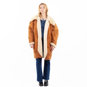 Puede incluir: Un abrigo largo de borrego marrón con interior y ribete color crema. El abrigo tiene un cuello ancho y bolsillos grandes. La persona de la imagen lleva vaqueros azules y zapatos negros.