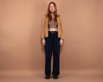 Puede incluir: Una mujer con una chaqueta de gamuza marrón, un top corto marrón, pantalones vaqueros azules y zapatos negros. Está de pie frente a un fondo marrón claro.