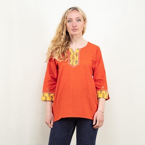 Könnte beinhalten: Eine orangefarbene Tunika mit Dreiviertelärmeln. Der Halsausschnitt und die Ärmelbündchen weisen ein gelbes und florales Muster auf. Die Tunika wird mit einer dunkelblauen Jeans kombiniert. Das Model hat lange blonde Haare.