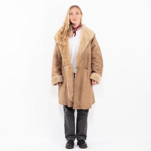 Könnte beinhalten: Ein langer, hellbrauner Mantel mit Shearling-Futter und einem großen Kragen. Der Mantel hat einen Knopfverschluss und wird mit dunkelgrauen Hosen und schwarzen Schuhen getragen. Die Manschetten und der Kragen sind mit einer helleren Shearling-Farbe gefüttert.