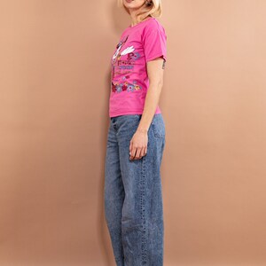 May include: A pink t-shirt with a colorful graphic print of a cartoon character. The shirt has a crew neck and short sleeves. The model is wearing blue jeans and black sneakers.