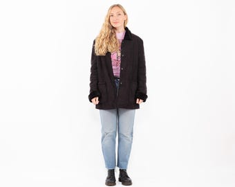 May include: A black quilted jacket with a faux fur collar and cuffs. The jacket has a button-up front and two pockets. The person is wearing a pink shirt and blue jeans. The jacket is a classic style, suitable for cooler weather.