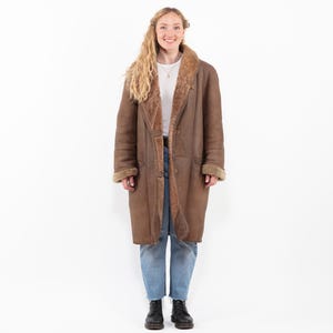 Puede incluir: Un abrigo largo de borrego marrón con cierre de botones. El abrigo tiene un cuello ancho y está forrado con borrego marrón más claro. Los puños también están forrados con borrego. La persona lleva vaqueros azules y botas negras.