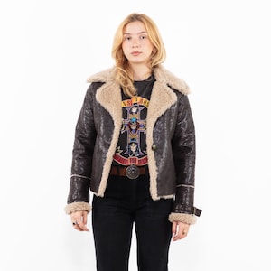 May include: A brown faux-leather jacket with a beige shearling collar and trim. The jacket has a button closure and a belt detail at the waist. The model is wearing a black t-shirt with a graphic print and black pants.