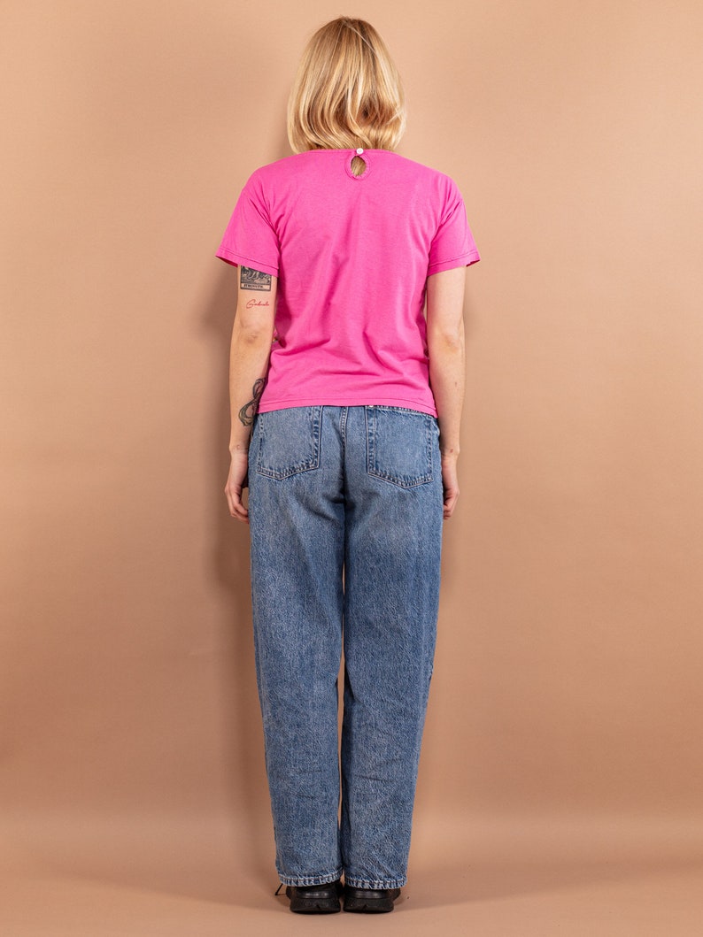 May include: A woman wearing a pink t-shirt and blue jeans. The t-shirt has a round neckline and short sleeves.