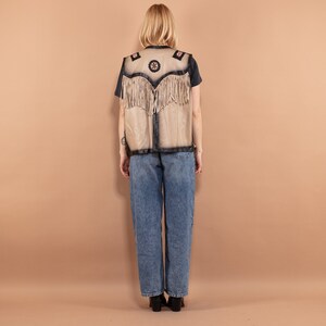 May include: A tan vest with black trim and fringe. The vest has decorative patches with floral and star designs. The person in the image is wearing a black t-shirt and blue jeans.