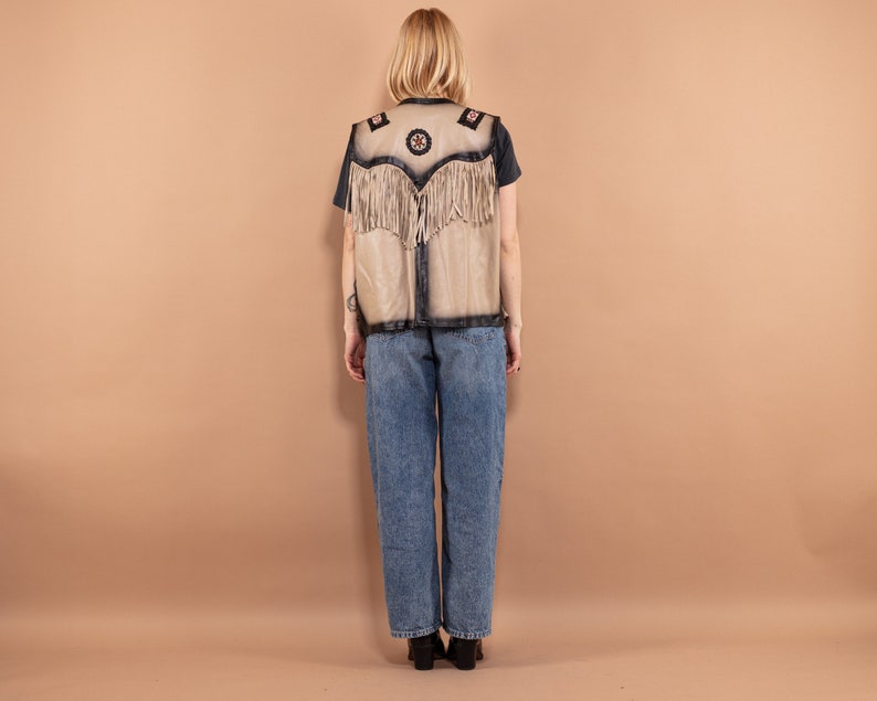 May include: A tan vest with black trim and fringe. The vest has decorative patches with floral and star designs. The person in the image is wearing a black t-shirt and blue jeans.
