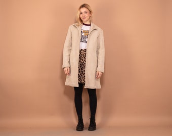 Puede incluir: Un abrigo beige de imitación de ante con cierre de cremallera, usado sobre una camiseta blanca y una falda con estampado de leopardo. El abrigo tiene cuello y llega hasta la pantorrilla. El conjunto se completa con leggings y botas negras.