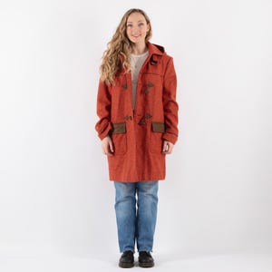 May include: A red wool coat with brown leather trim and toggle fastenings. The coat has a hood and pockets. The model is wearing blue jeans and brown shoes.