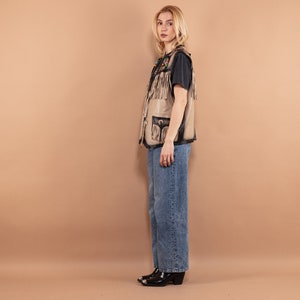 May include: A tan leather vest with fringe and black accents. The vest has pockets and decorative stitching. The person is wearing a black t-shirt and blue jeans. The boots are black with silver tips.