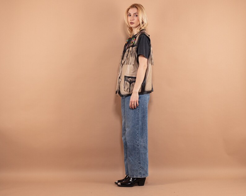 May include: A tan leather vest with fringe and black accents. The vest has pockets and decorative stitching. The person is wearing a black t-shirt and blue jeans. The boots are black with silver tips.