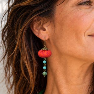 Puede incluir: Pendientes colgantes que presentan una pequeña cuenta redonda de tela roja texturizada en la parte superior, seguida de dos pequeñas cuentas de color turquesa y una cuenta verde oscuro, unidas por piezas y ganchos de metal color bronce.