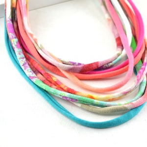 May include: A multi-strand necklace made of colorful fabric strips. The necklace is made of a variety of colors, including pink, orange, green, and blue. The fabric is patterned with floral designs.