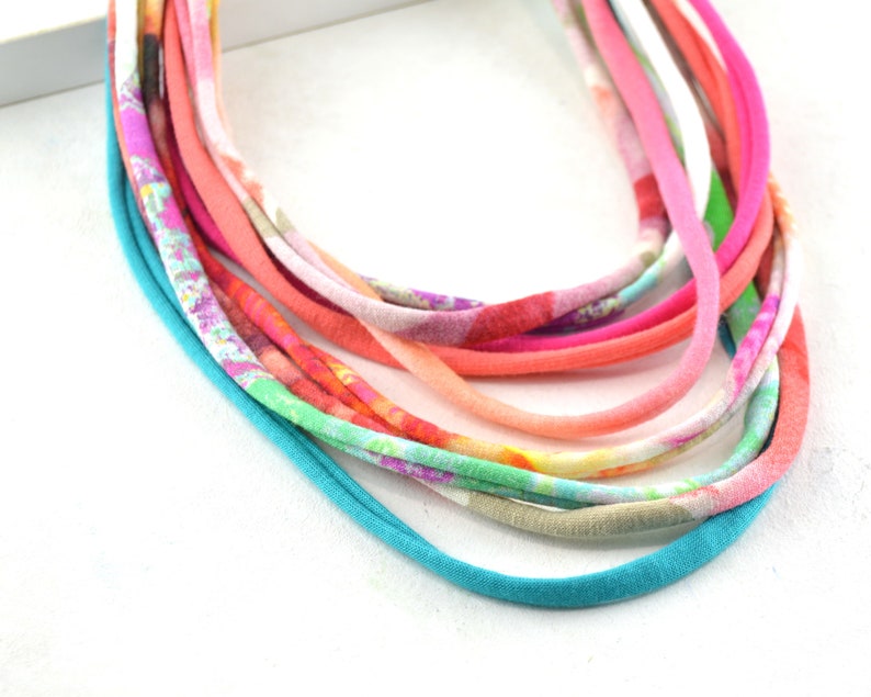 May include: A multi-strand necklace made of colorful fabric strips. The necklace is made of a variety of colors, including pink, orange, green, and blue. The fabric is patterned with floral designs.