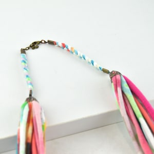 May include: A colorful braided necklace with a pink, white, and blue tassel. The necklace is made of fabric and has a metal clasp.
