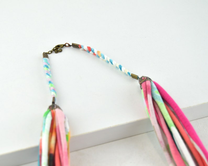 May include: A colorful braided necklace with a pink, white, and blue tassel. The necklace is made of fabric and has a metal clasp.
