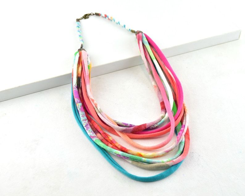 May include: Multi-strand necklace made of colorful fabric strips. The necklace is layered with different colors and patterns, including pink, orange, green, and blue.