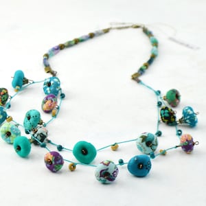 Puede incluir: Un collar de múltiples hilos con cuentas de color turquesa y con estampado floral. El collar está hecho de tela y cuentas.