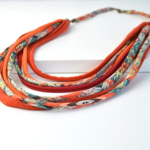 Puede incluir: Un collar de tela de varias hebras con capas de cordones textiles suaves en naranja, rojo oscuro y estampados. Las secciones estampadas muestran un diseño geométrico en tonos azul verdoso, crema y marrón, asegurado con un cierre de metal.