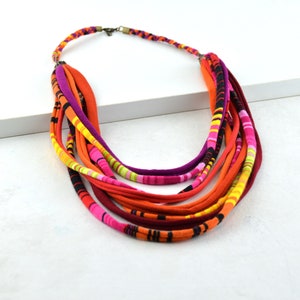 May include: Multi-colored fabric necklace with a layered design. The necklace is made of several strands of fabric in shades of orange, pink, yellow, and black.