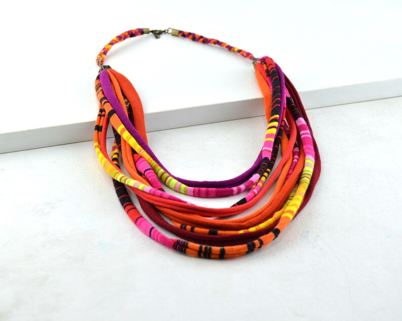 May include: Multi-colored fabric necklace with a layered design. The necklace is made of several strands of fabric in shades of orange, pink, yellow, and black.