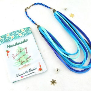May include: A handmade necklace made of multiple strands of blue fabric. The necklace is displayed on a white surface with a small gift bag that says "Handmade Something special for you" and "Daniela Barbieri".