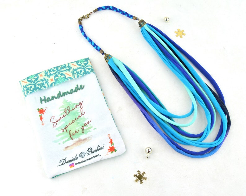 May include: A handmade necklace made of multiple strands of blue fabric. The necklace is displayed on a white surface with a small gift bag that says "Handmade Something special for you" and "Daniela Barbieri".