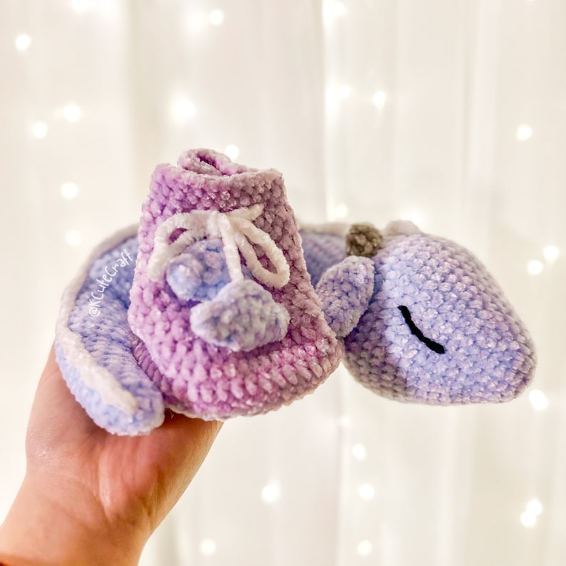 K&ouml;nnte beinhalten: Ein Paar geh&auml;kelte Babyschuhe in Lila- und Blaut&ouml;nen. Die Schuhe haben ein wei&szlig;es Band, das um sie herum gebunden ist, und einen kleinen blauen Bommel an jedem Zeh.