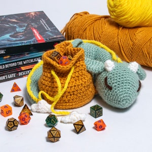 K&ouml;nnte beinhalten: Ein geh&auml;kelter Drachen-W&uuml;rfelbeutel mit gelbem Kordelzug und einem gr&uuml;n-blauen Drachenkopf. Der Beutel ist mit bunten W&uuml;rfeln gef&uuml;llt und liegt auf einer wei&szlig;en Oberfl&auml;che.