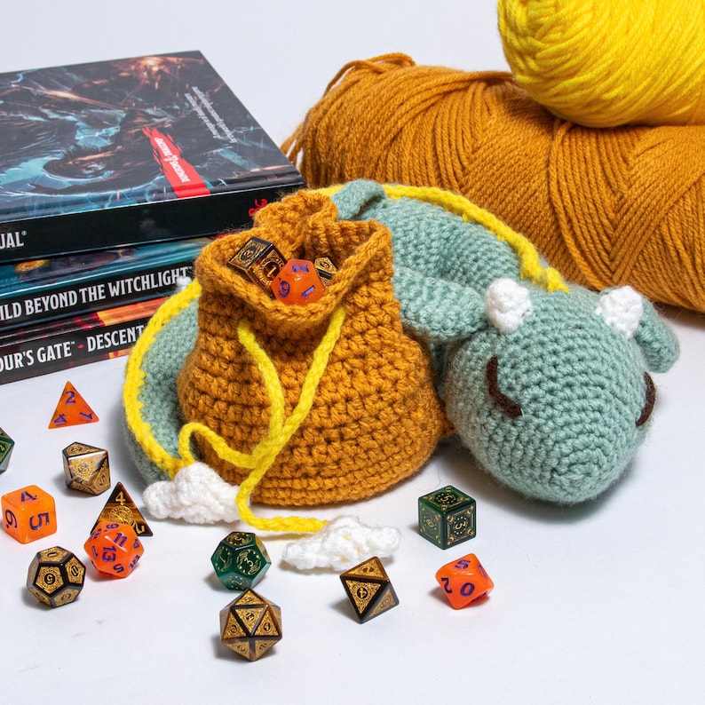 K&ouml;nnte beinhalten: Ein geh&auml;kelter Drachen-W&uuml;rfelbeutel mit gelbem Kordelzug und einem gr&uuml;n-blauen Drachenkopf. Der Beutel ist mit bunten W&uuml;rfeln gef&uuml;llt und liegt auf einer wei&szlig;en Oberfl&auml;che.