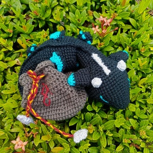 K&ouml;nnte beinhalten: Ein schwarzer und blauer geh&auml;kelter Drache mit wei&szlig;en Augen, zusammengerollt mit einer braunen geh&auml;kelten Tasche mit einem roten und gelben Kordelzug.
