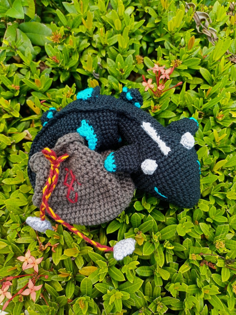 K&ouml;nnte beinhalten: Ein schwarzer und blauer geh&auml;kelter Drache mit wei&szlig;en Augen, zusammengerollt mit einer braunen geh&auml;kelten Tasche mit einem roten und gelben Kordelzug.